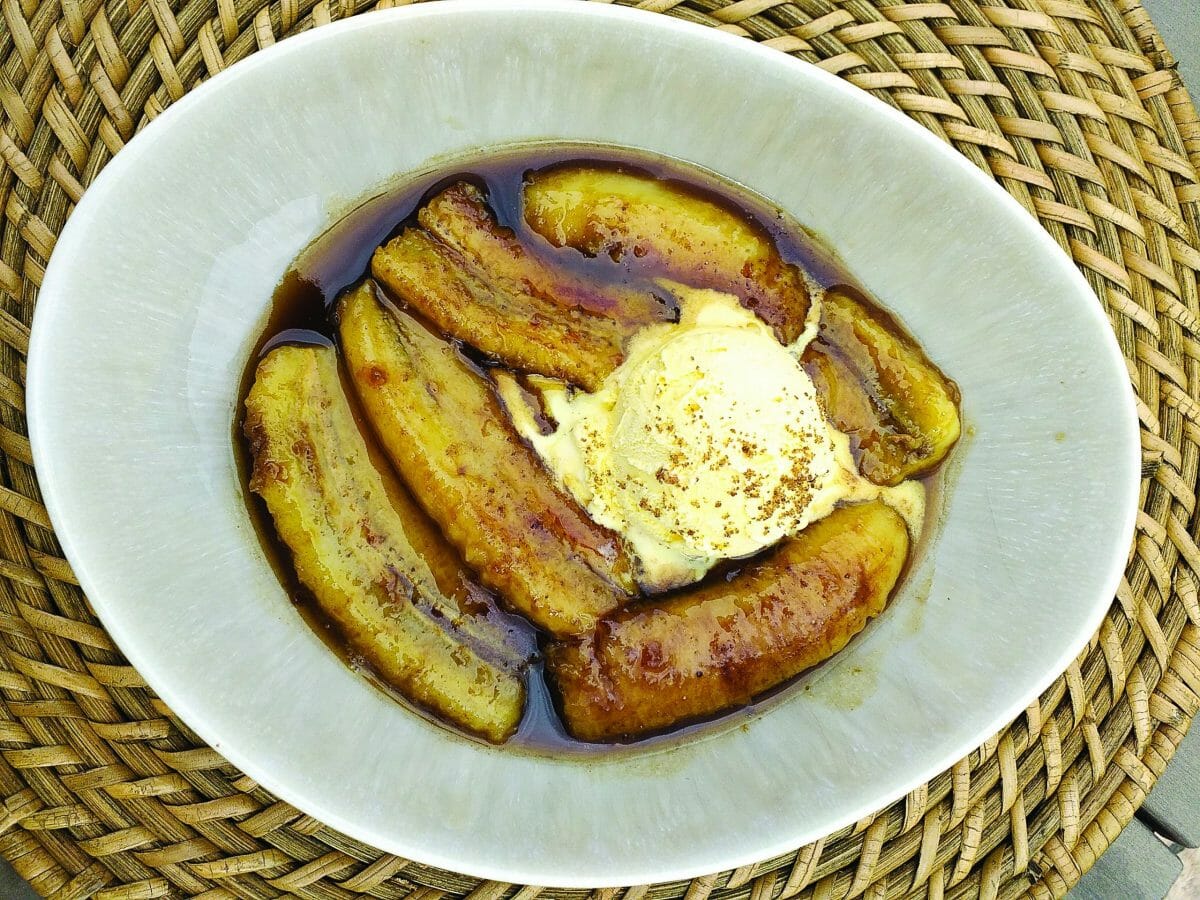 Assiette creuse grise sur un sous plat en paille, 6 morceaux de bananes cuites qui baignent dans la sauce, une boule de glace vanille au centre.