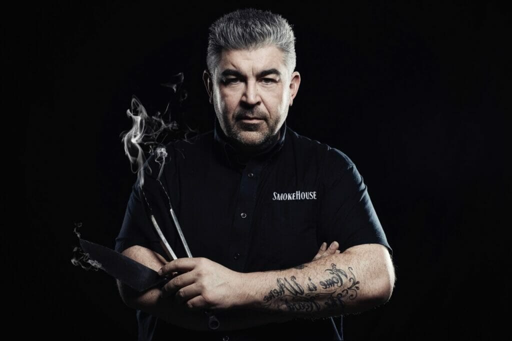 Une photo de Chef Rohan sur fond noir avec des pinces de BBQ qui fument