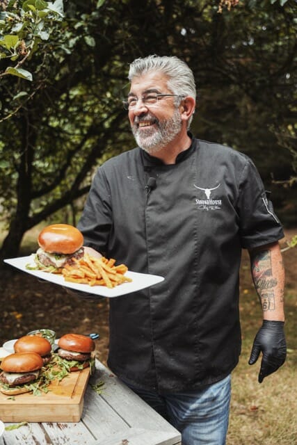 Chef Rohan souriant en tablier noir, une assiette avec un burger et des frites à la main