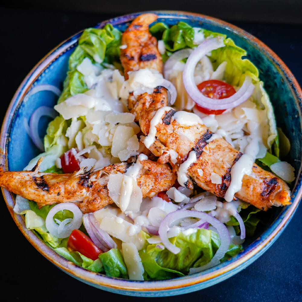 Salade, filets de poulet grillés, parmesan en copeaux, oignons rouges et sauce César.