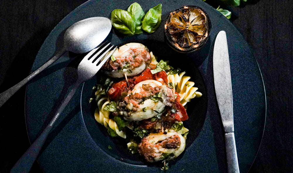assiette noire avec sur un lit de torteloni, des encornets farcis. Un demi citron grillé et des couverts argentés